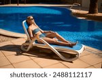 Side view of a woman lying on a poolside sun lounger with her arms at her side, sunbathing, tropical landscape in the background. Summer holidays, holiday enjoyment, travel concept