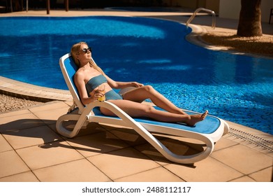 Side view of a woman lying on a poolside sun lounger with her arms at her side, sunbathing, tropical landscape in the background. Summer holidays, holiday enjoyment, travel concept - Powered by Shutterstock