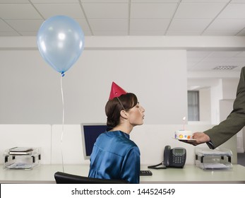 Side view of a woman in party hat blowing birthday candle on cake in office - Powered by Shutterstock