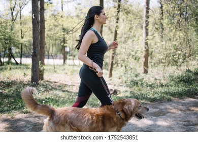 Side View Of Woman In Sportswear Walking With Dog In Park. Sport Girl Walking Her Dog In Nature During Sunny Day.