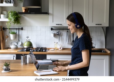 Side View Woman Standing In Domestic Cozy Kitchen Wear Headphones Having Distance Communication, Looks At Laptop Use Video Call App, Studying Working Remotely At Home. Virtual Meeting, E-learn Concept