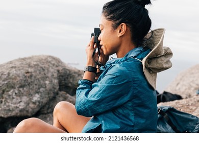 Side View Of A Woman Taking Photographs On Her Film Camera While Sitting On A Rock During A Mountain Hike