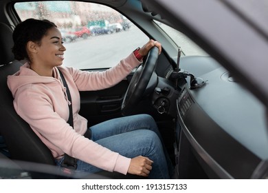 Side view of woman taxi driver sits in transport, holding steering wheel and looking on road. Young smiling african american female driving car in town in raining, near phone with gps map, copy space