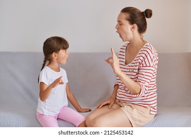 Side View Of Woman Teacher Wearing Striped Shirt And Shorts Sitting On Sofa With Little Girl, Preschooler Kid Practicing Correct Pronunciation With A Female Speech Therapist.