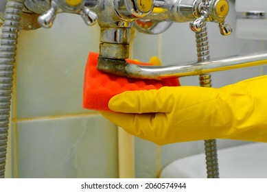 Side view of woman's hand in rubber glove with sponge wiping rusty, old chrome faucet. Close-up housewife cleaning plumbing fixtures from dirt in bathtub indoors. Household cleaning, bathroom washing.