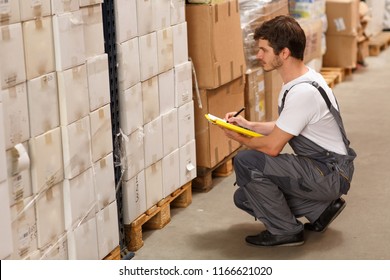 Side view of worker crouching down, looking at goods and writing something in yellow clipboard. Man wearing uniform and white t shirt. Background of warehouse with lots of boxes.