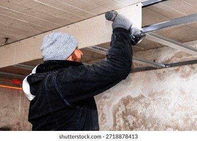 Side view of a worker fastening ceiling framing with an electric drill. Home renovation indoors, manual labor, tools and construction process in progress.