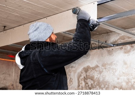 Side view of a worker fastening ceiling framing with an electric drill. Home renovation indoors, manual labor, tools and construction process in progress.