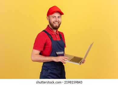 Side View Of Worker Man Wearing Blue Overalls, Red T-shirt And Cap Working On Laptop, Expressing Positive Emotions, Looking At Camera. Indoor Studio Shot Isolated On Yellow Background.