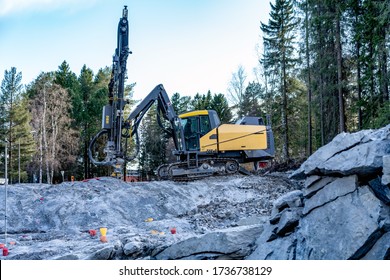 Side view at yellow drilling machine which stands on granite rock and boring holes for dynamite at rock blasting area - further rock exploding, new road construction. Evening shadows, green forest