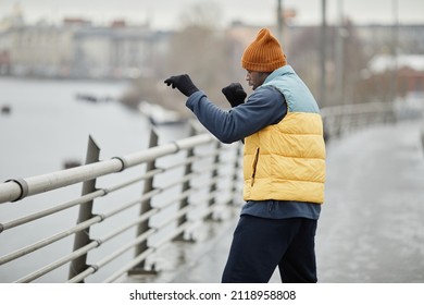 Side view of young active blackman in sportswear practicing boxing exercise while standing in front of riverside in the morning