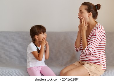 Side View Of Young Adult Caucasian Language Therapist Teaching Little Kid Sounds Pronunciation, Working On Speech Defects Or Difficulties With Small Child Girl Indoors While Sitting On Sofa.