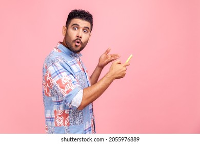 Side View Of Young Adult Man Pointing At Mobile Phone With Shocked Astonished Expression, Looking At Camera With Big Eyes And Open Mouth, Indoor Studio Shot Isolated On Pink Background.