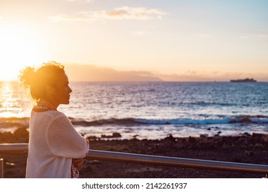 Side view of young adult woman admiring and enjoying golden sunset at the beach looking at the sea. Female people and nature outdoor leisure activity. Thoughtful lady and travel life - Powered by Shutterstock