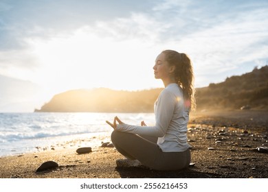 Side view of a young adult woman meditating in lotus yoga pose. A calm female yogi practices yoga and meditations among the sea and mountains at the sunset. Do yoga and spiritual practice outdoors.
