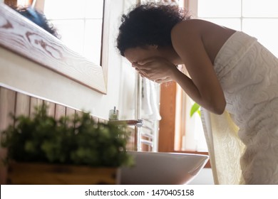 Side view young african american woman wrapped in towel after showering, bending over sink, washing off mask or peeling, cleaning her face with pure running water. Daily hygiene skincare routine. - Powered by Shutterstock