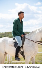 Side view of young African American male looking away while sitting on back of white horse on ranch