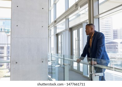 Side View Of Young African American Businessman Standing On A Mezzanine Leaning Handrail In The Atrium Of A Bright Modern Business Building. Modern Corporate Start Up New Business Concept With