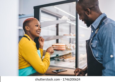 Side view of young African American man in dark apron choosing pastry and speaking with stylish black woman in yellow shirt and turquoise apron pointing on cake  - Powered by Shutterstock