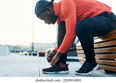 Side View Of Young African American Runner In Black Leggins And Cap And Red Sport Jacket Bending Over Sneaker While Tying Shoelace