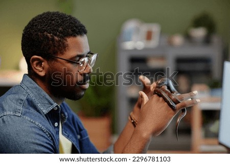 Side view of young African American man looking at snake creeping over his hands during alternative therapy session in home environment