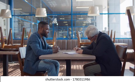 Side View Of Young African Entrepreneur Mature Bank Manager Signing Contract In Empty Cafe. Small Business Owner Taking Loan For Startup Development And Opening Restaurant