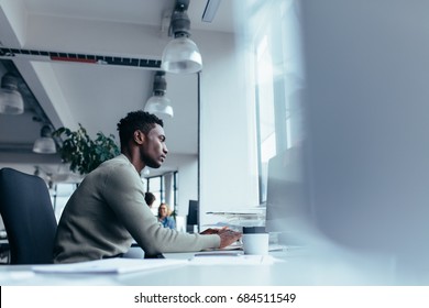 Side View Of Young African Man Working In Office. Male Executive Sitting At His Desk And Working On Desktop Computer.