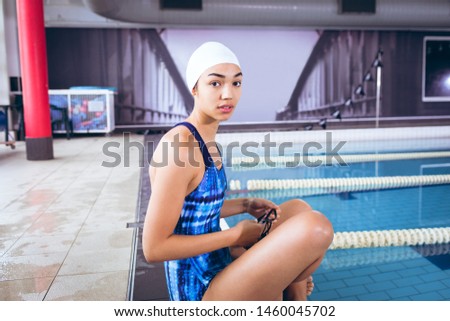 Side view of a young African-American woman wearing a swimsuit and swimming cap sitting by an olympic sized pool inside a stadium