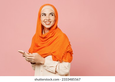 Side view young arabian asian muslim woman wear orange abaya hijab hold in hand use mobile cell phone look aside isolated on plain pink background. People uae middle eastern islam religious concept - Powered by Shutterstock