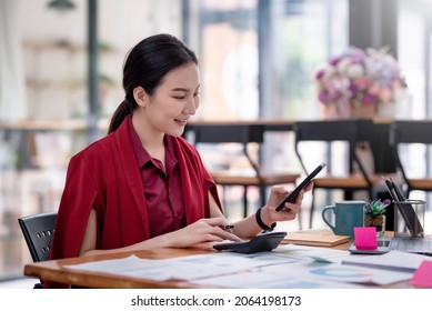 Side view of young Asian businesswoman holding a smartphone using a calculator to calculate work graph papers placed on the table. - Powered by Shutterstock