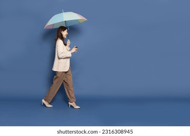 Side view of young Asian businesswoman walking and holding hot coffee cup and umbrella isolated on blue background, In the rainy season concept - Powered by Shutterstock