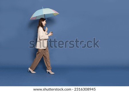Side view of young Asian businesswoman walking and holding hot coffee cup and umbrella isolated on blue background, In the rainy season concept