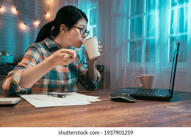 side view of young asian japanese elegant housewife counting finance in dark night home kitchen and eating tasty instant noodles. lady drink ramen soup as bedtime snack while looking at laptop screen