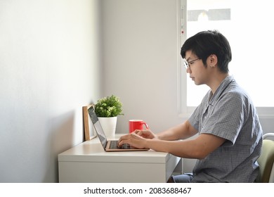 Side View Young Asian Man In Pajamas Checking Email On Laptop Computer At Home.