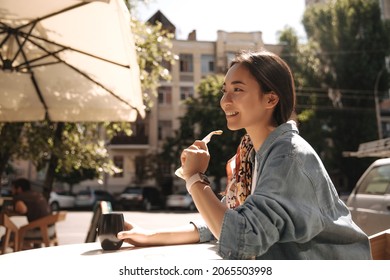 Side view of young asian stylish woman in denim jacket drinking coffee outdoors in cafe. Dark long hair gathered in ponytail, looking in front of her. People and free time concept