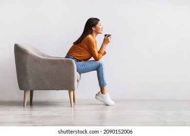 Side view of young Asian woman sitting in armchair, drinking hot aromatic coffee, relaxing against white studio wall, free space. Lovely millennial lady enjoying warm beverage on lazy day - Shutterstock ID 2090152006
