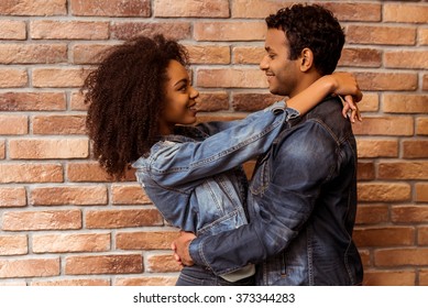 Side view of young attractive Afro-American couple hugging, looking in camera and smiling while standing against brick wall