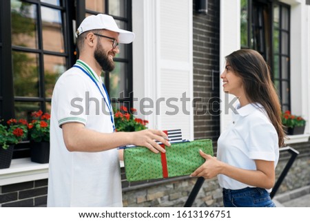 Side view of young bearded deliveryboy in white cap giving two gift boxes to woman. Pretty smiling brunette female in casual outfit receiving order and folder to sign near house. Delivery concept.
