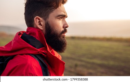 Side view of young bearded male hiker in warm red jacket looking away while standing against blurred green field during trekking in autumn countryside