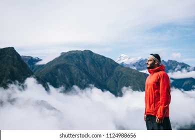 Side view of young bearded man dressed in active wear for trip standing on background with high mountains covered mist during trekking wanderlust.Male traveler admiring beauty of natural environment