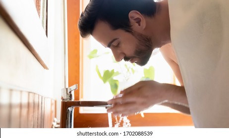 Side view young bearded man washing face with running tap water, bend over sink in bathroom. Millennial handsome guy doing morning skincare hygiene routine after showering, head shot close up view.