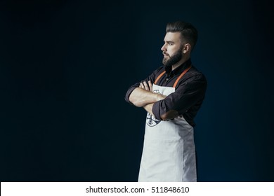 Side View. A Young Bearded Man, Dressed In A Dark Brown Shirt And Bright Apron, Standing With His Arms Folder. On Background Dark Blue Wall.