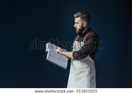 Side view. A young bearded man, dressed in a dark brown shirt and bright apron, holding a thick book with recipes and reading. In the background dark blue wall.