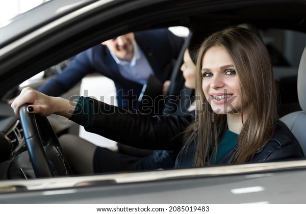 Side view of young beautiful woman sitting
inside car and holding hand on steering wheel. She smiling and
talking with manager of car dealership. Car agent representing
inside of automobile.