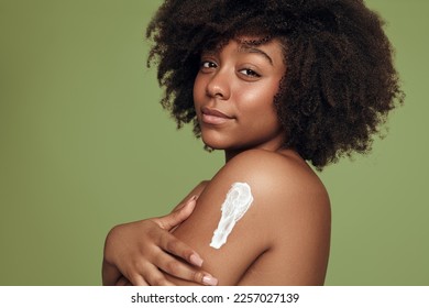 Side view of young black female with Afro hairstyle applying body lotion on arm and looking at camera during spa session against green background - Shutterstock ID 2257027139