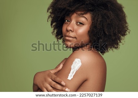 Side view of young black female with Afro hairstyle applying body lotion on arm and looking at camera during spa session against green background