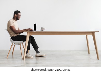 Side view of young black guy in casual wear using laptop, sitting at desk, working online from home office, copy space. Millennial African American man freelancing, having remote job