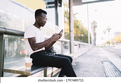 Side View Of Young Black Man In Eyeglasses With Black Backpack Sitting On Bench At Bus Stop With Takeaway Coffee And Using Smartphone While Waiting For Bus On Warm Evening