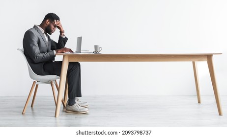 Side view of young black office employee feeling tired of working overtime, sitting at desk with laptop against white studio wall, copy space. Exhausted businessman having problem with online job - Shutterstock ID 2093798737