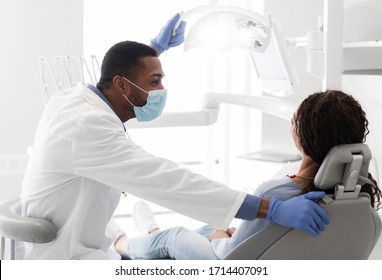 Side View Of Young Black Stomatologist Talking To His Female Patient Sitting In Dentist Chair